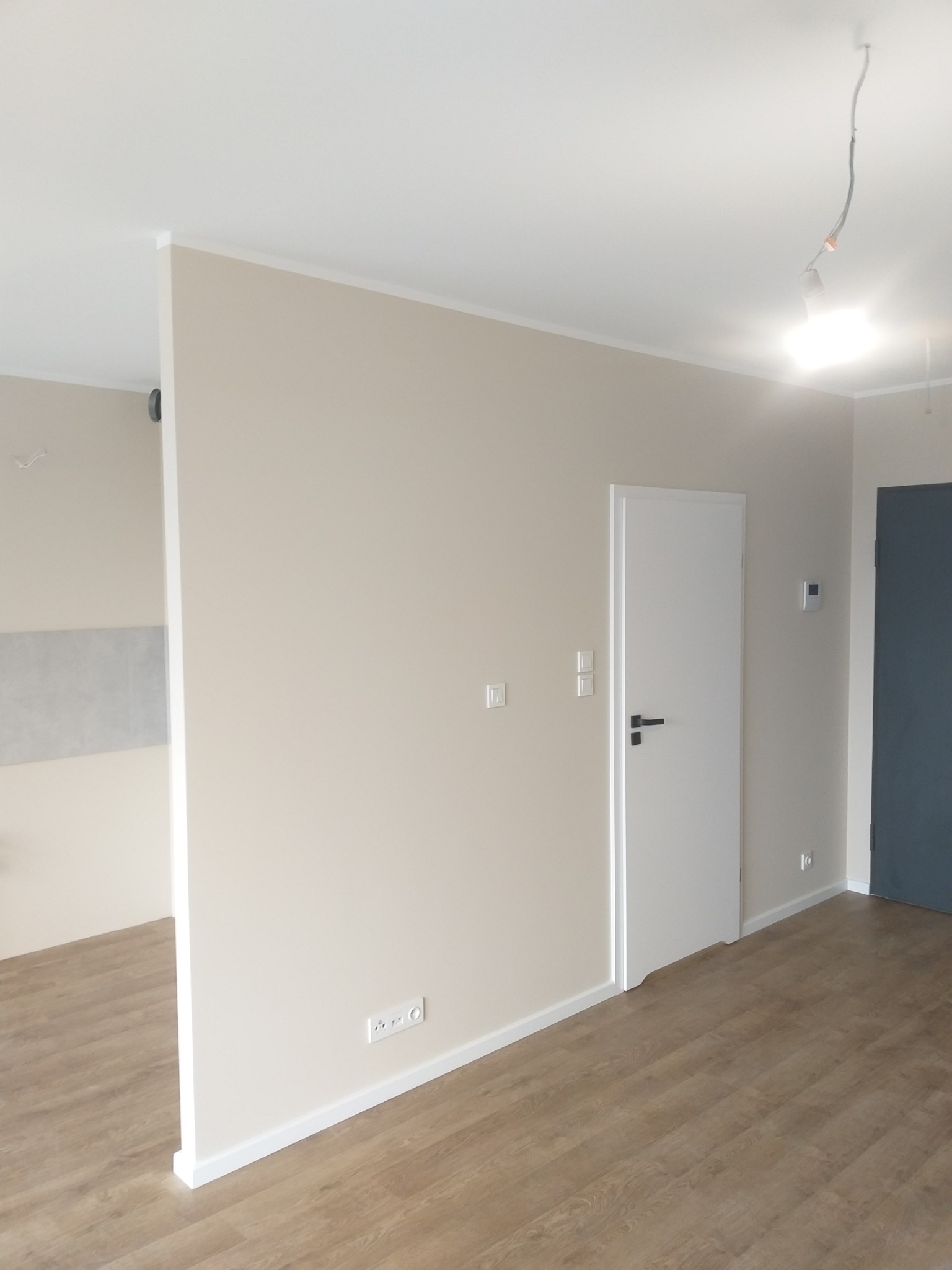 Wnętrze po remoncie: ściany w odcieniach beżu, drewnopodobna podłoga, białe drzwi z czarną klamką, widoczne gniazdka elektryczne i włączniki światła.
