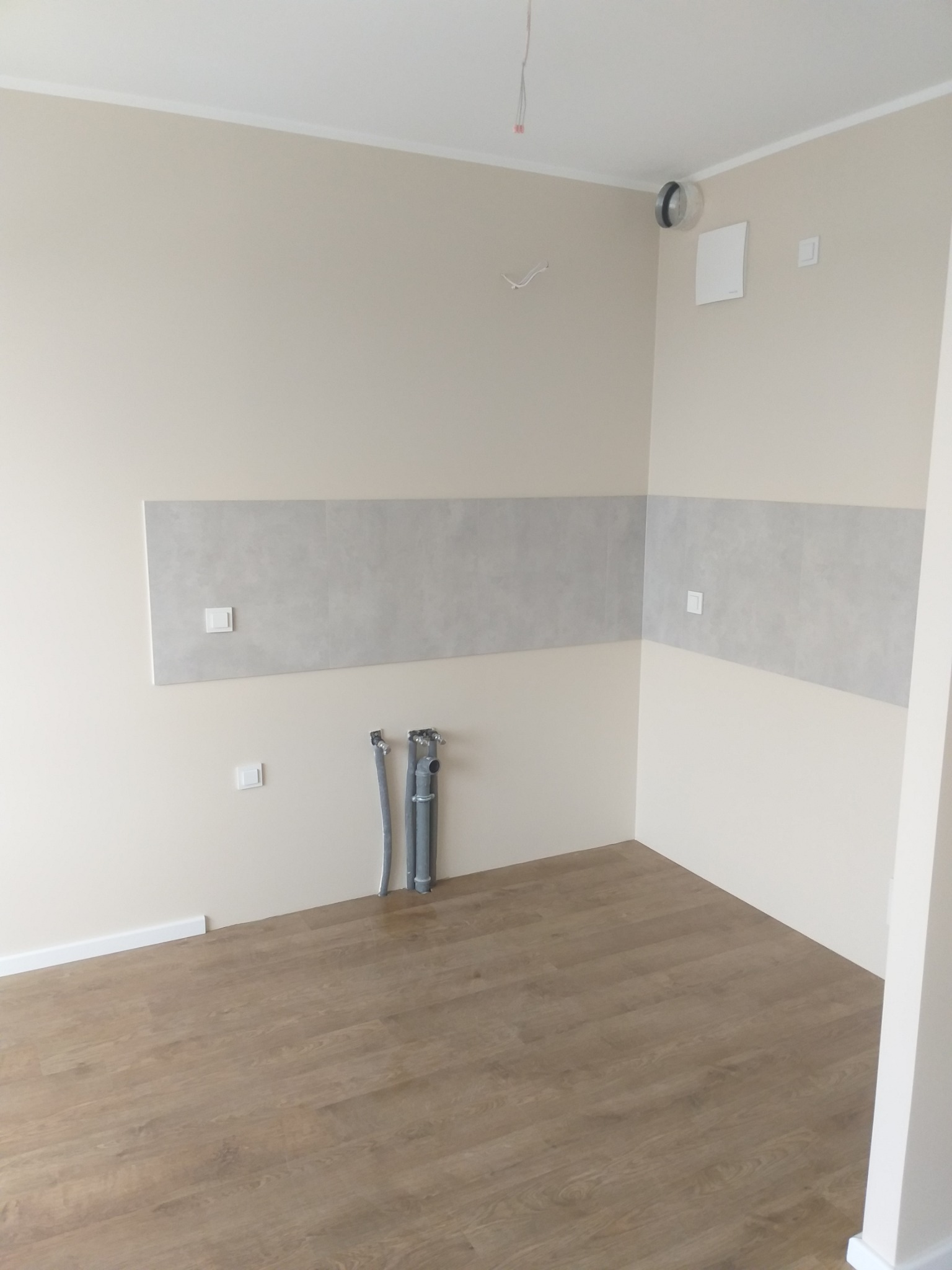 Wnętrze pomieszczenia w trakcie wykańczania, widoczne szare płytki na ścianie, drewnopodobna podłoga, wystające rury instalacji wodnej i elektryczne przewody. Ściany pomalowane na jasny beż.