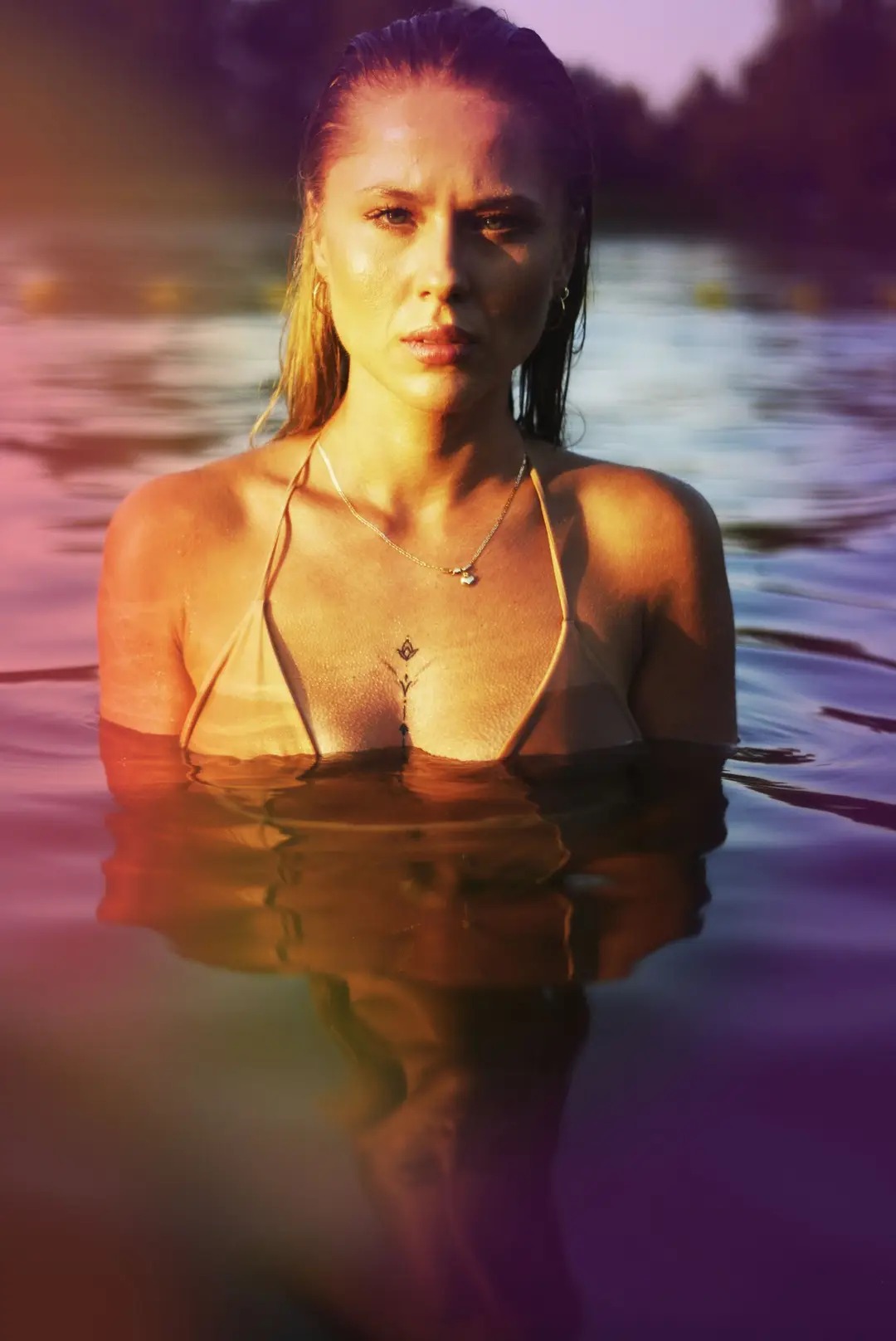 Portret kobiety zanurzonej w wodzie po ramiona, w bikini, z mokrymi włosami i delikatnym tatuażem na dekolcie, patrzącej wprost w obiektyw, z refleksami światła na wodzie i skórze.