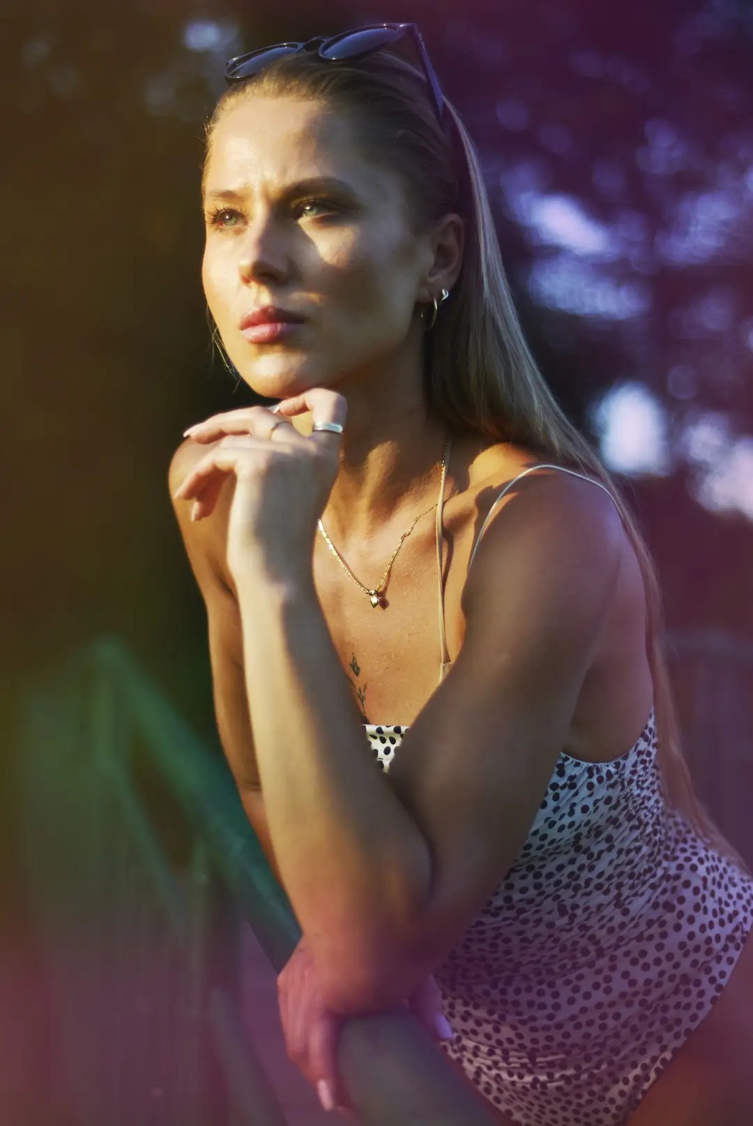 Portret młodej kobiety z blond włosami, opierającej podbródek na dłoni, z okularami przeciwsłonecznymi na głowie i delikatnym tatuażem na dekolcie, w letnim stroju w kropki.