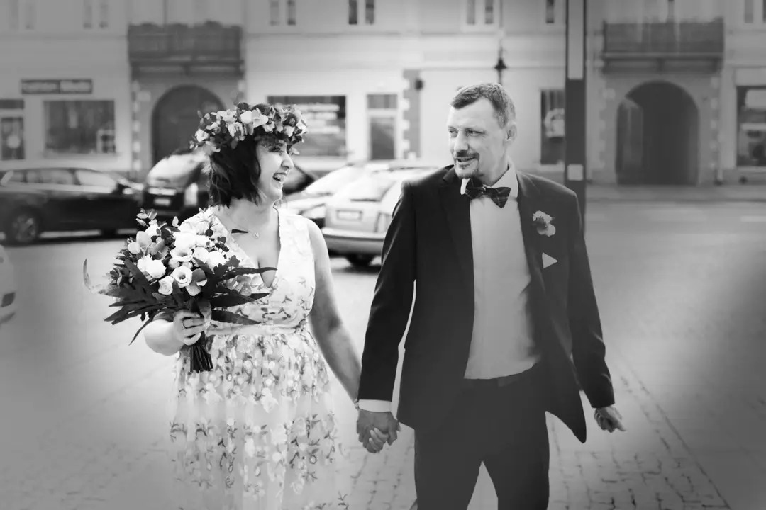Czarno-białe zdjęcie uśmiechniętej pary młodej trzymającej się za ręce na tle miejskiej ulicy z budynkami i zaparkowanymi samochodami. Panna młoda ma na głowie wianek, a w ręku bukiet białych...