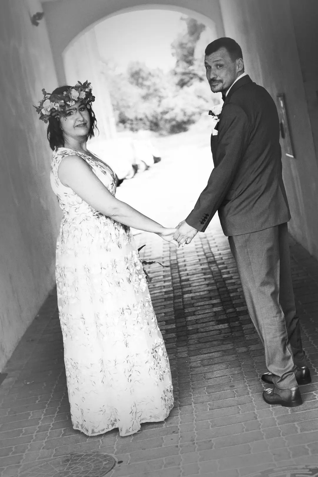 Czarno-białe zdjęcie pary trzymającej się za ręce w tunelu; kobieta w sukience w kwiaty i wianku, mężczyzna w garniturze.