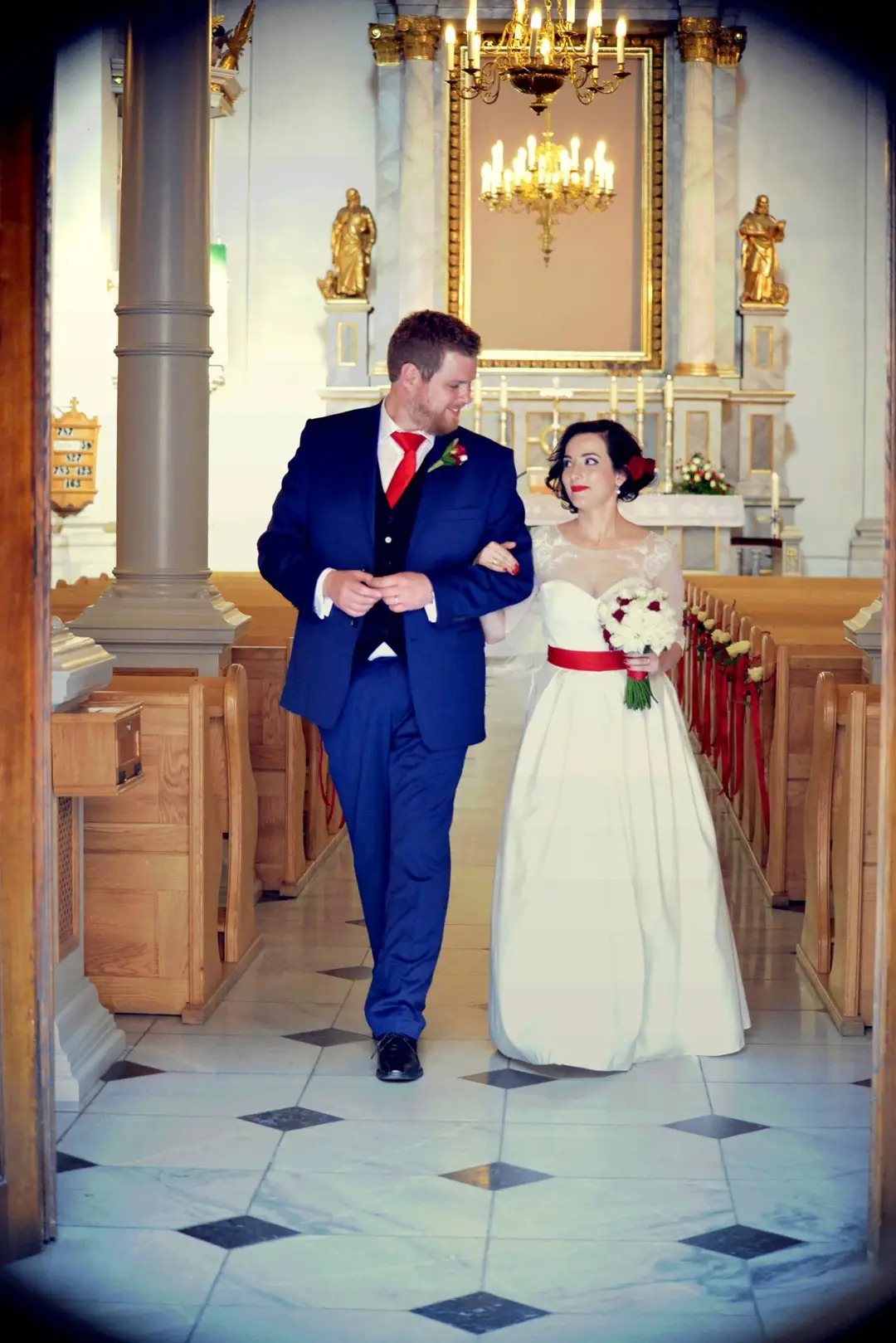 Para młoda w kościele: Pan młody w niebieskim garniturze z czerwoną muchą, panna młoda w białej sukni z czerwonym paskiem, trzyma bukiet. W tle ołtarz i złote dekoracje.