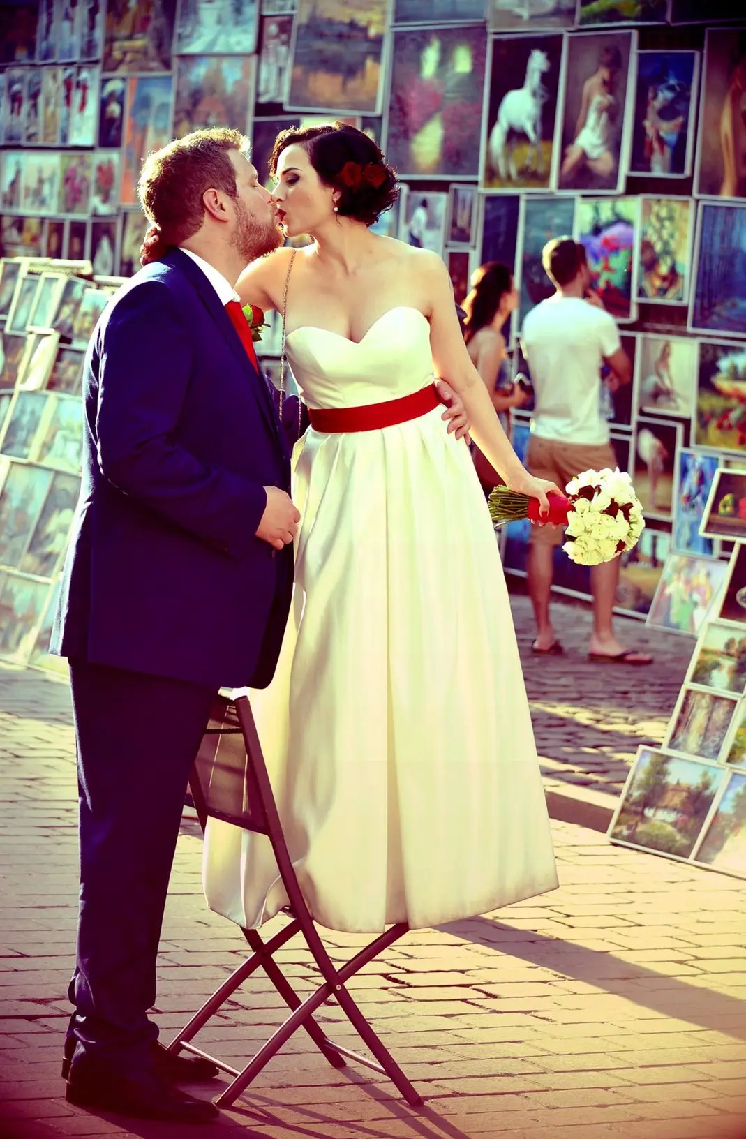 Pocałunek pary młodej na tle straganu z obrazami: panna młoda w białej sukni z czerwoną szarfą, pan młody w granatowym garniturze, kwiaty we włosach, bukiet w dłoni.
