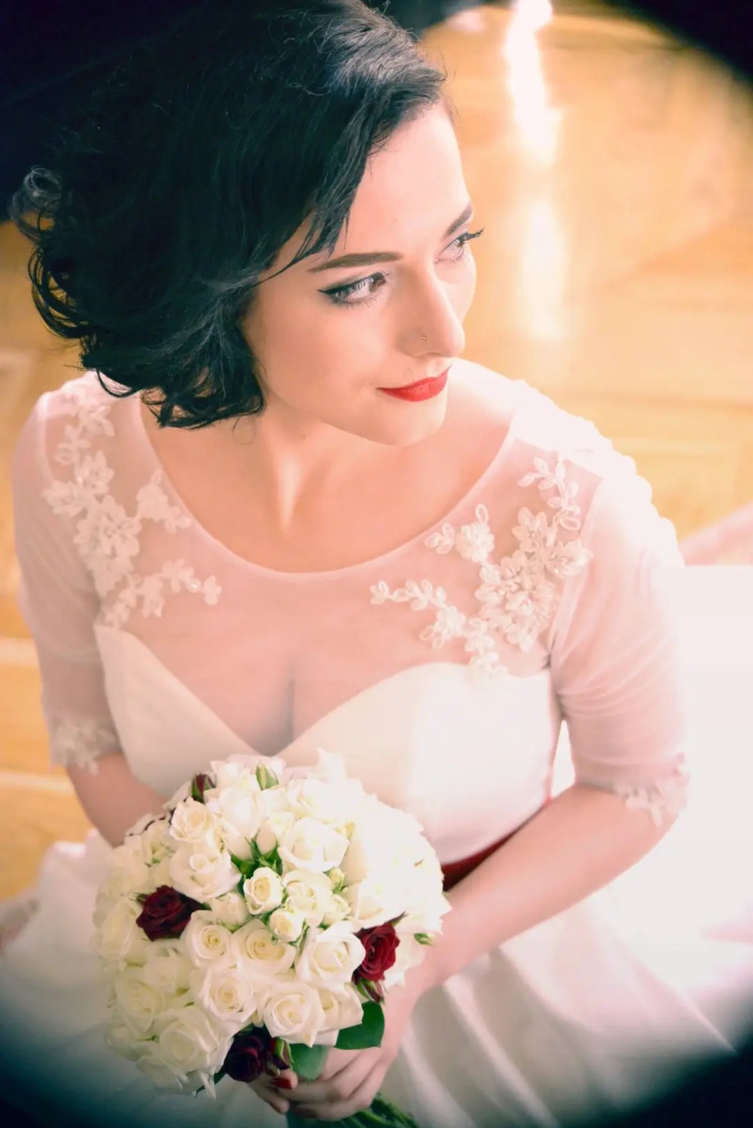 Panna młoda z ciemnymi włosami, w sukni ślubnej z koronkową górą, trzyma bukiet białych róż z akcentami burgundu, spogląda w bok na rozmytym tle parkietu.