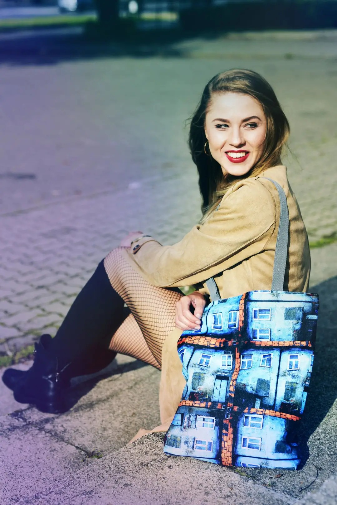 Uśmiechnięta młoda kobieta w beżowym płaszczu i rajstopach w siatkę siedzi na betonowym podłożu, trzymając torebkę z nadrukiem przedstawiającym budynki z niebieskimi ścianami i pomarańczowymi...