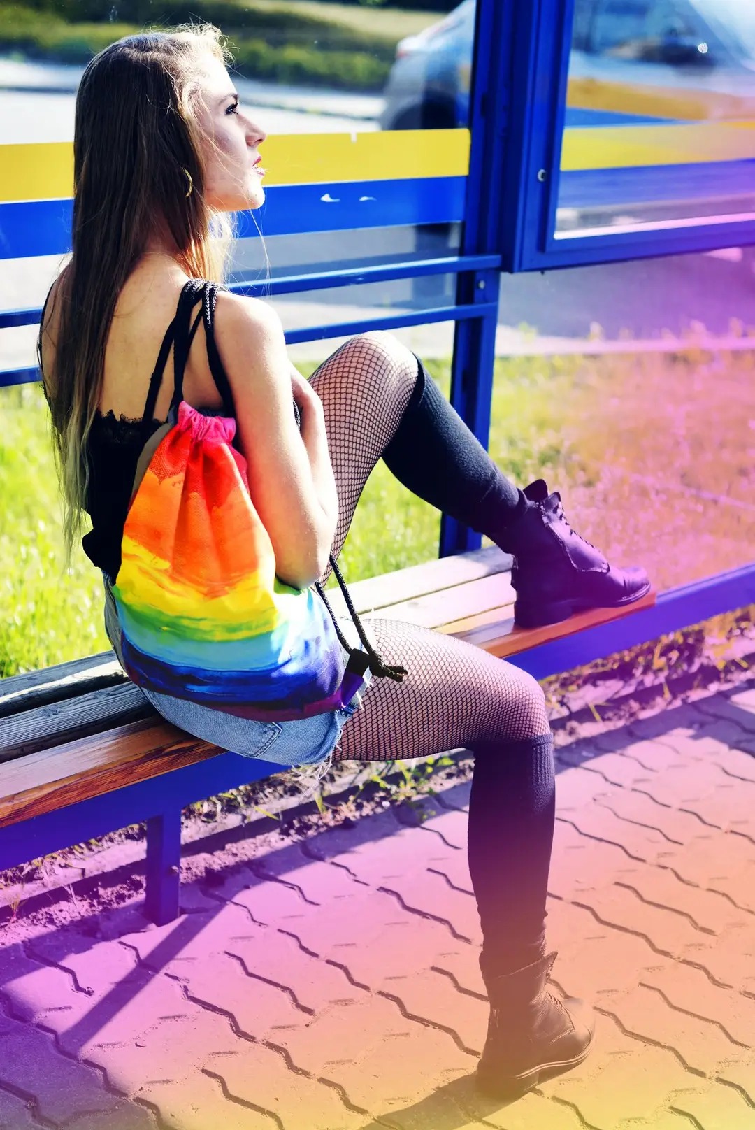 Młoda kobieta z plecakiem w tęczowe pasy siedzi na przystanku autobusowym, ubrana w kabaretki i czarne buty sznurowane.