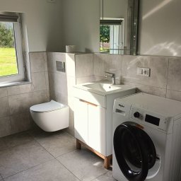 Usługi remontowo budowlane Tomasz Lewicz - Nowoczesna łazienka z podwieszaną toaletą, umywalką z szafką na drewnianych nóżkach, pralką Whirlpool i lustrem w ramie, wykończona szarymi płytkami.