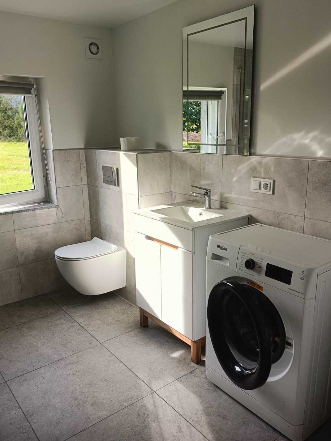 Nowoczesna łazienka z podwieszaną toaletą, umywalką z szafką na drewnianych nóżkach, pralką Whirlpool i lustrem w ramie, wykończona szarymi płytkami.