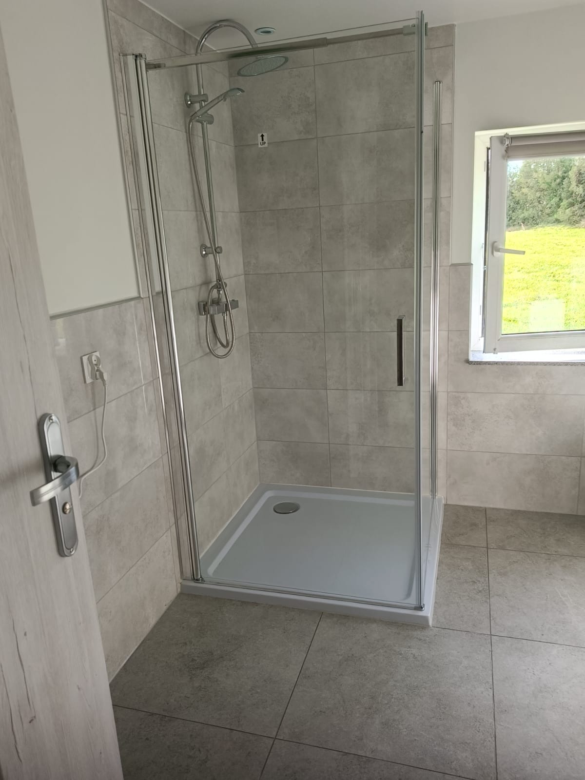 Nowoczesna kabina prysznicowa z szarymi płytkami, widok z boku, z widocznym oknem z zielonym krajobrazem w tle, minimalistyczny design.