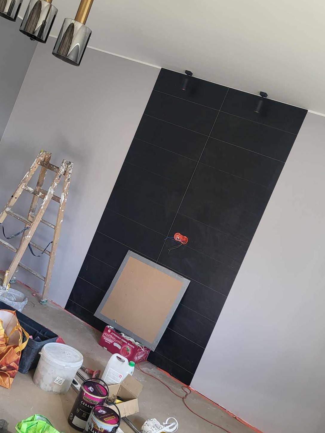 Częściowo wykończona ściana z czarnymi płytkami, widoczne puszki elektryczne, drabina malarska, farby i akcesoria malarskie na podłodze, przygotowania do dalszych prac wykończeniowych.