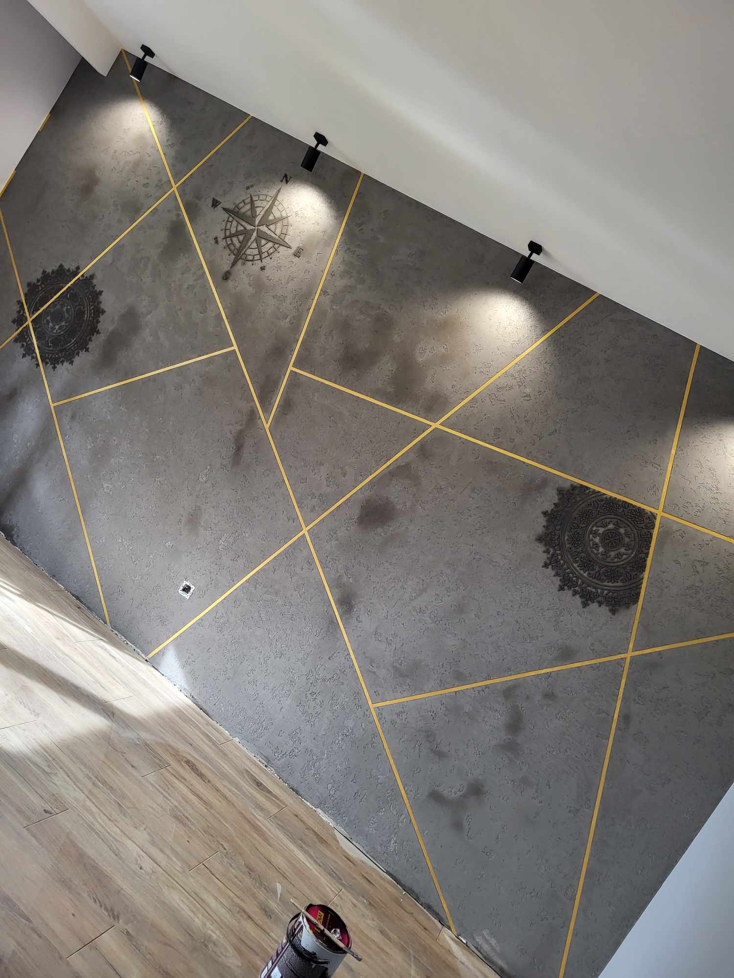 Dekoracyjna szara ściana z geometrycznymi złotymi liniami, rozetami i kompasem, oświetlona światłem punktowym, widok na podłogę z paneli.