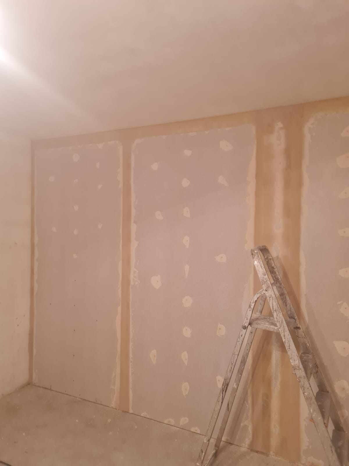 Wnętrze pokoju z płytami kartonowo-gipsowymi, widoczne łączenia płyt wypełnione masą szpachlową i miejsca po wkrętach, drabina aluminiowa oparta o ścianę, surowy stan przed dalszym wykończeniem.