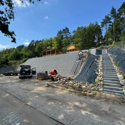 Krystian Płotka - Wzmocnienie skarpy z wykorzystaniem ażurowych płyt betonowych, schodów i siatki zabezpieczającej, widoczne narzędzia i betoniarka, w tle drewniane altany na szczycie skarpy.