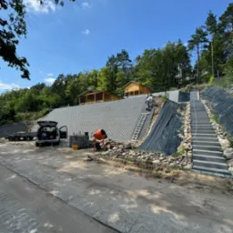 Wzmocnienie skarpy z wykorzystaniem ażurowych płyt betonowych, schodów i siatki zabezpieczającej, widoczne narzędzia i betoniarka, w tle drewniane altany na szczycie skarpy.