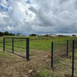 Nowo postawione, ciemnozielone ogrodzenie panelowe z otwartą furtką na działce z widokiem na budujące się domy i pochmurne niebo.