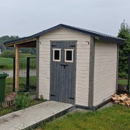 Drewniany domek narzędziowy z dwuspadowym dachem, pomalowany na beżowo i szaro, z małym zadaszeniem i ścieżką z płyt betonowych na trawniku.