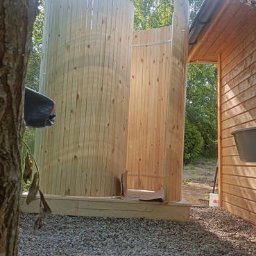 Drewniana sauna beczka w trakcie budowy, widoczna z zewnątrz, z elementami konstrukcyjnymi i otoczeniem naturalnym. Częściowo zmontowana konstrukcja.