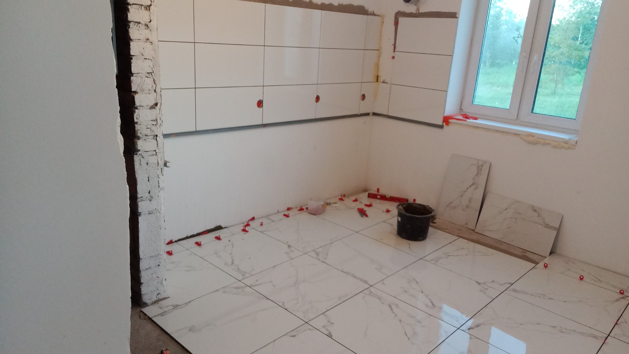 Układanie białych płytek z szarym marmurkowym wzorem na podłodze i ścianie w trakcie remontu, widoczne czerwone krzyżyki poziomujące, wiadro i poziomica, surowy fragment ściany z cegły.