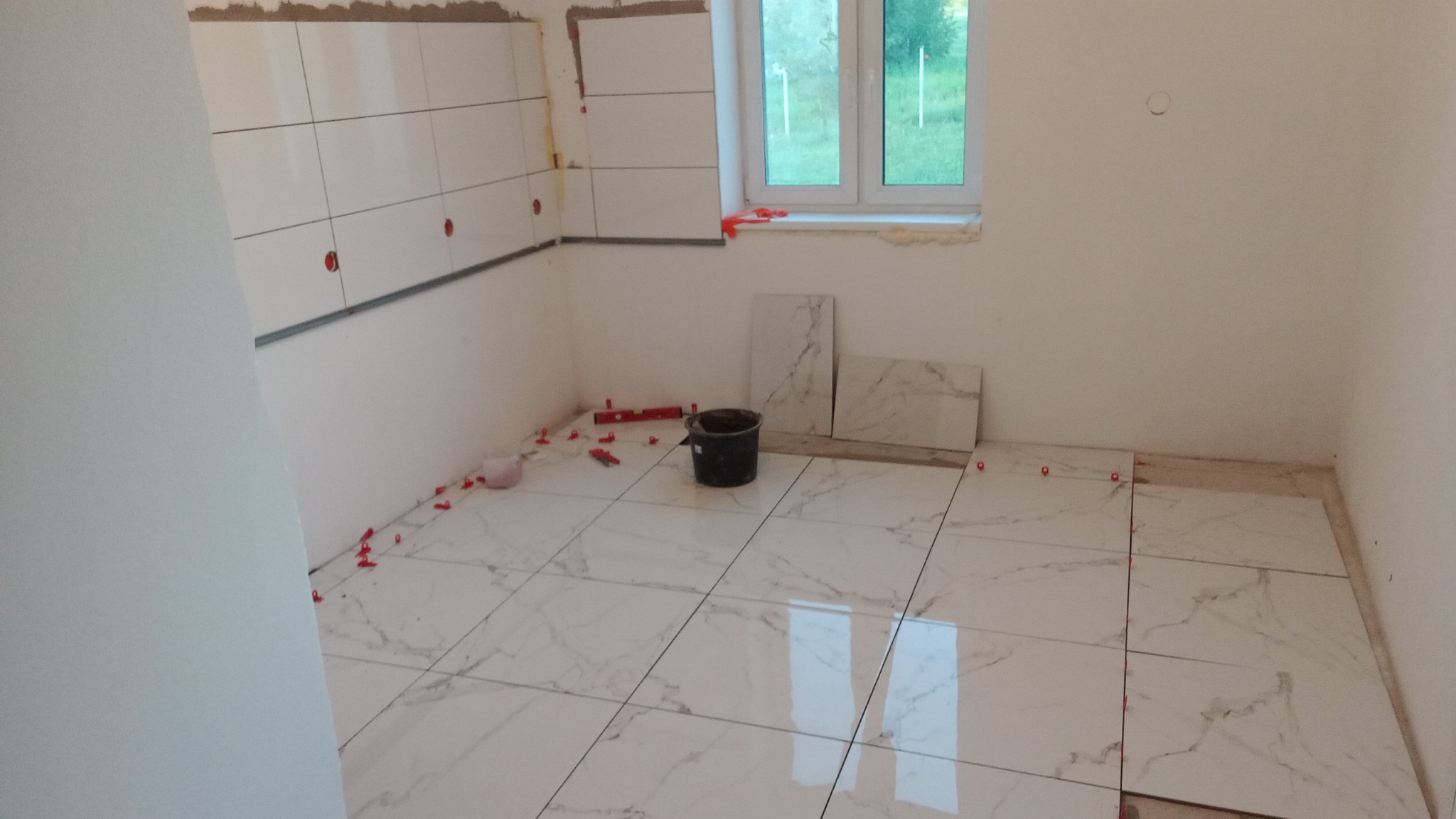 Układanie białych płytek imitujących marmur na podłodze i ścianie w trakcie remontu łazienki, widoczne krzyżyki dystansowe, poziomica i czarne wiadro.