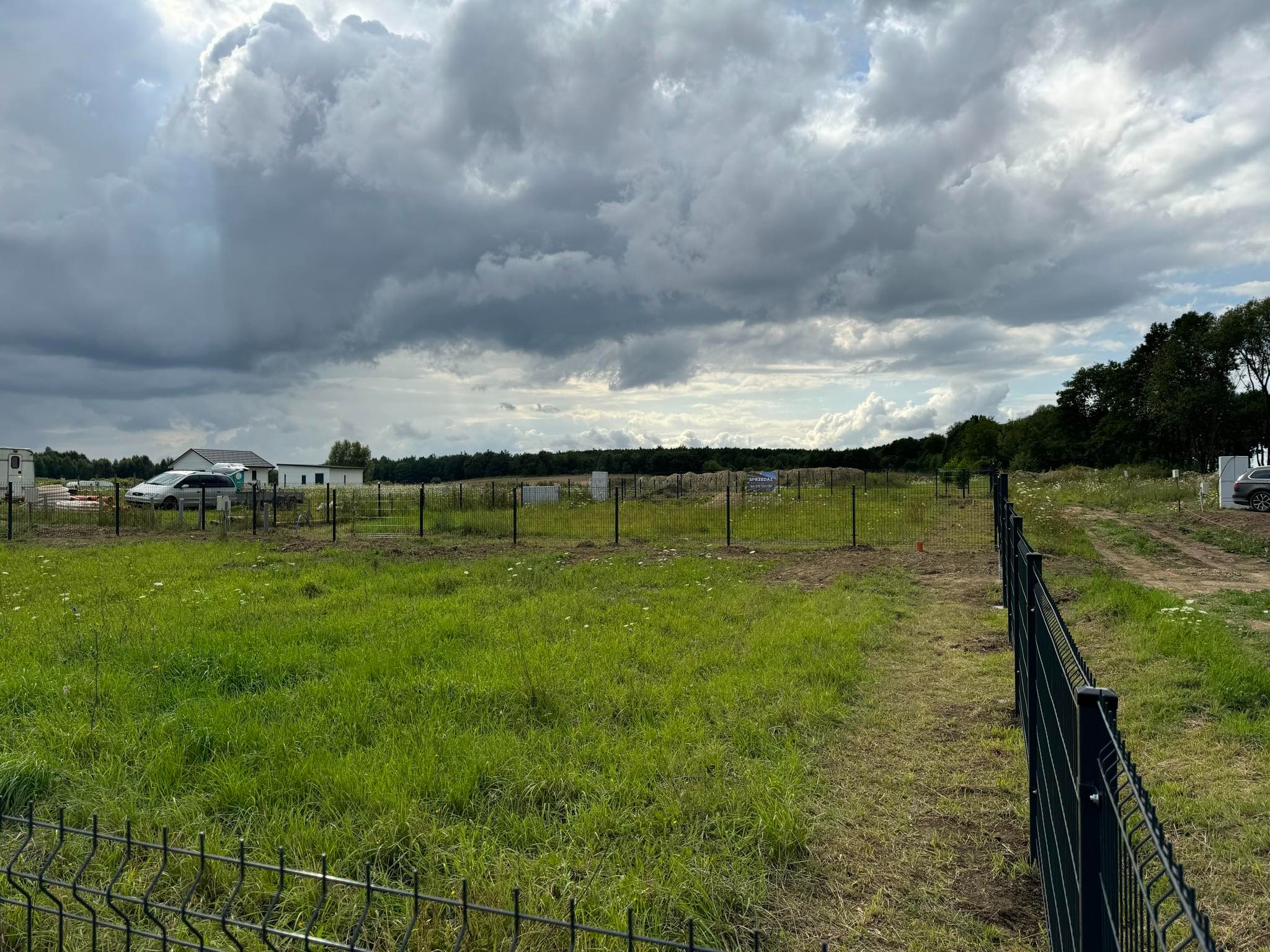 Działka budowlana z nowo postawionym, czarnym ogrodzeniem panelowym na tle pochmurnego nieba i zabudowań gospodarczych w oddali.
