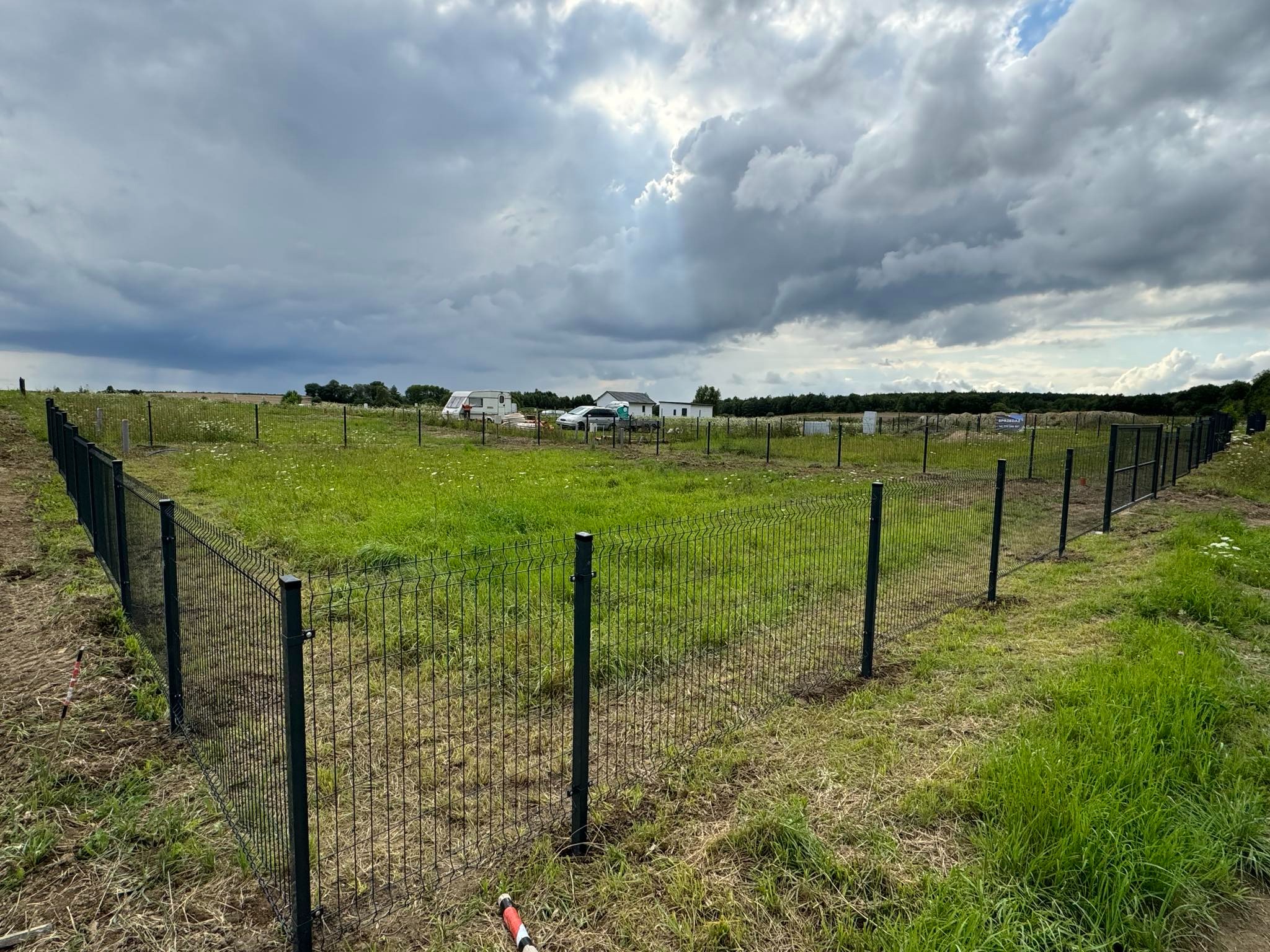 Nowo postawione, ciemne ogrodzenie panelowe na zielonej, otwartej przestrzeni z widocznymi w oddali zabudowaniami i pochmurnym niebem.