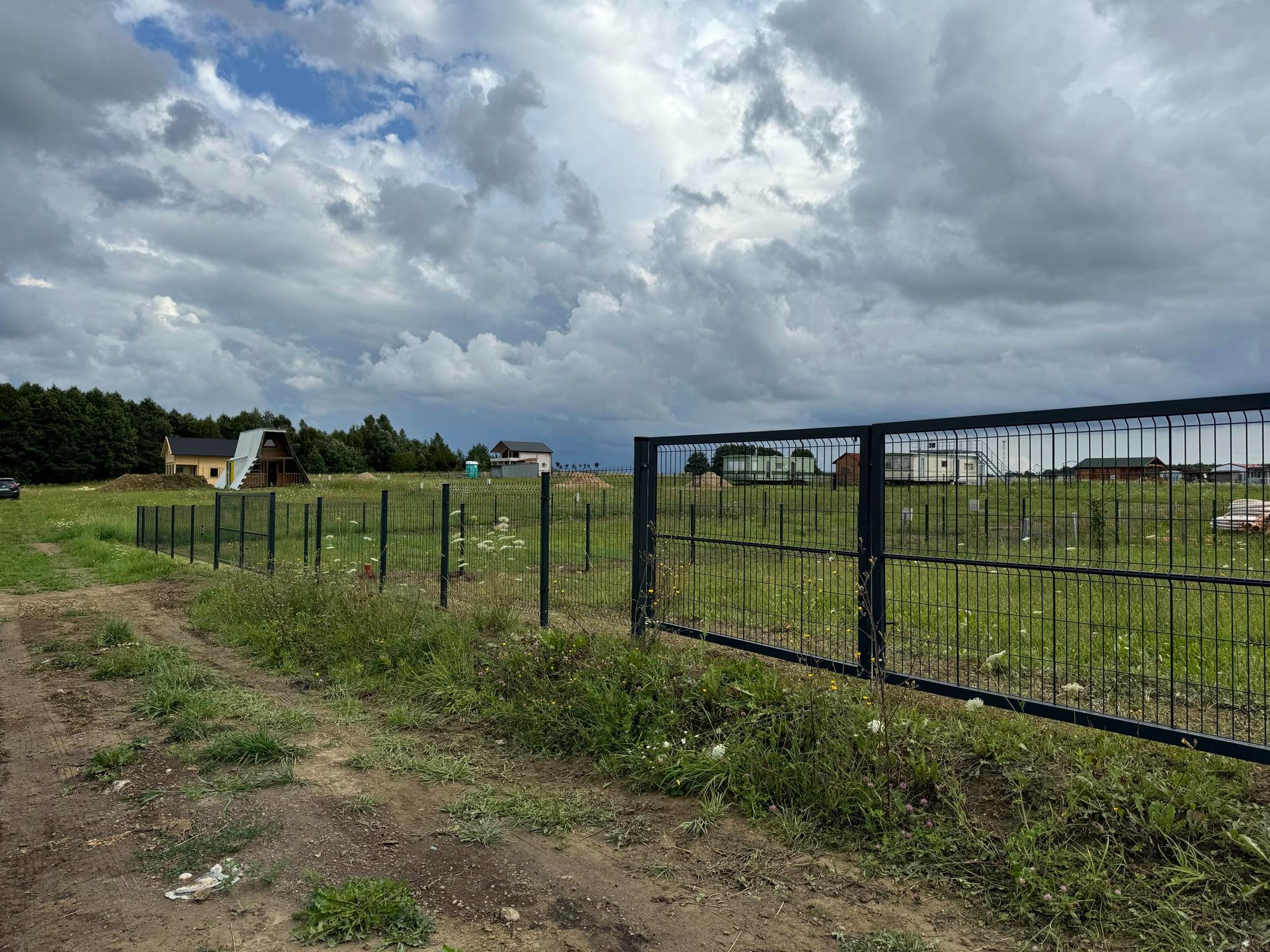 Nowoczesne ogrodzenie panelowe z metalu, otaczające posesję z widocznymi domami w tle, pochmurne niebo.