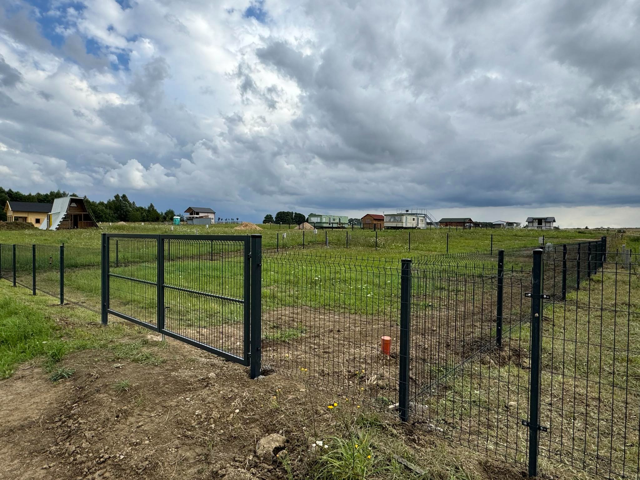 Nowo postawione, ciemnozielone ogrodzenie panelowe z otwartą furtką na działce z widokiem na budujące się domy i pochmurne niebo.