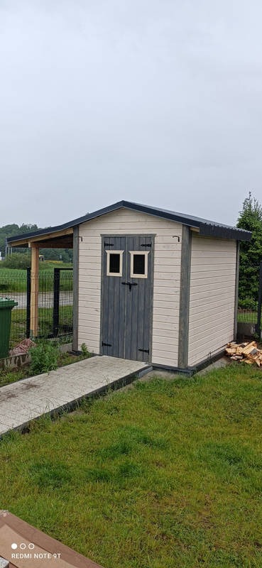 Drewniany domek narzędziowy z dwuspadowym dachem, pomalowany na beżowo i szaro, z małym zadaszeniem i ścieżką z płyt betonowych na trawniku.