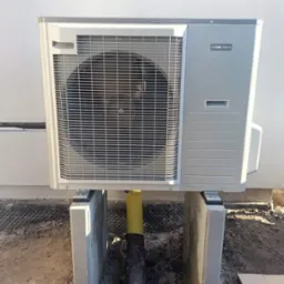 Zewnętrzna jednostka klimatyzacji NIBE AMS10 zamontowana na betonowych wspornikach, z ż&oacute;łtą rurą odprowadzającą skropliny do wykopanego dołu.