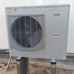 Szara zewnętrzna jednostka klimatyzacji NIBE AMS10, zamontowana na betonowych słupkach, widoczna na tle białej ściany i gruntu z folią fundamentową.