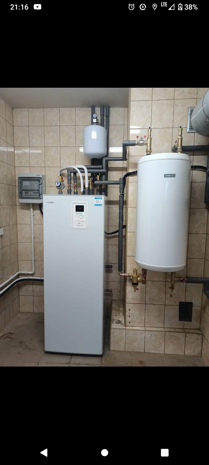 Biały piec grzewczy marki Nibe z podłączonymi rurami i zbiornikiem, zamontowany w pomieszczeniu z beżowymi płytkami.