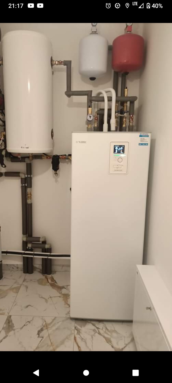 Biały piec gazowy Nibe z wyświetlaczem i podłączonymi rurami oraz zbiornikami, zamontowany w pomieszczeniu z marmurową podłogą.