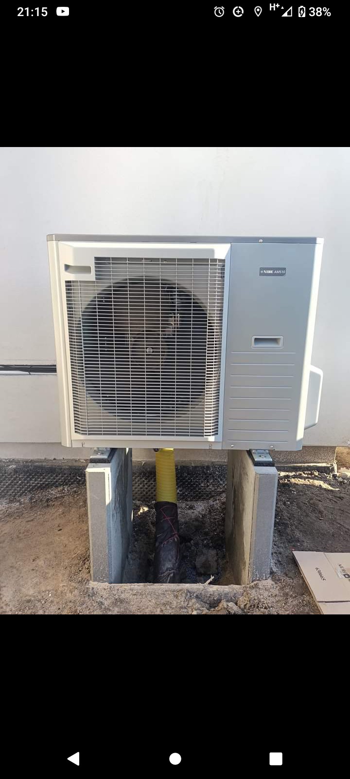 Zewnętrzna jednostka klimatyzacji NIBE AMS10 zamontowana na betonowych wspornikach, z żółtą rurą odprowadzającą skropliny do wykopanego dołu.