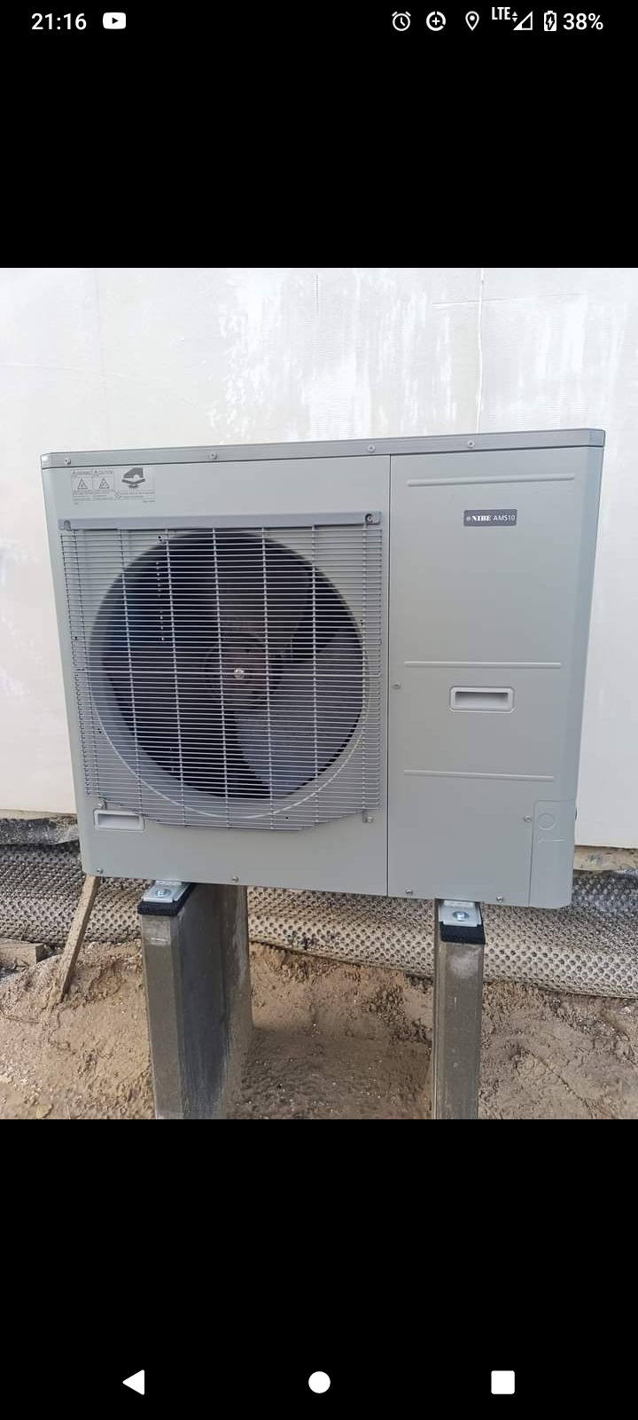 Szara zewnętrzna jednostka klimatyzacji NIBE AMS10, zamontowana na betonowych słupkach, widoczna na tle białej ściany i gruntu z folią fundamentową.