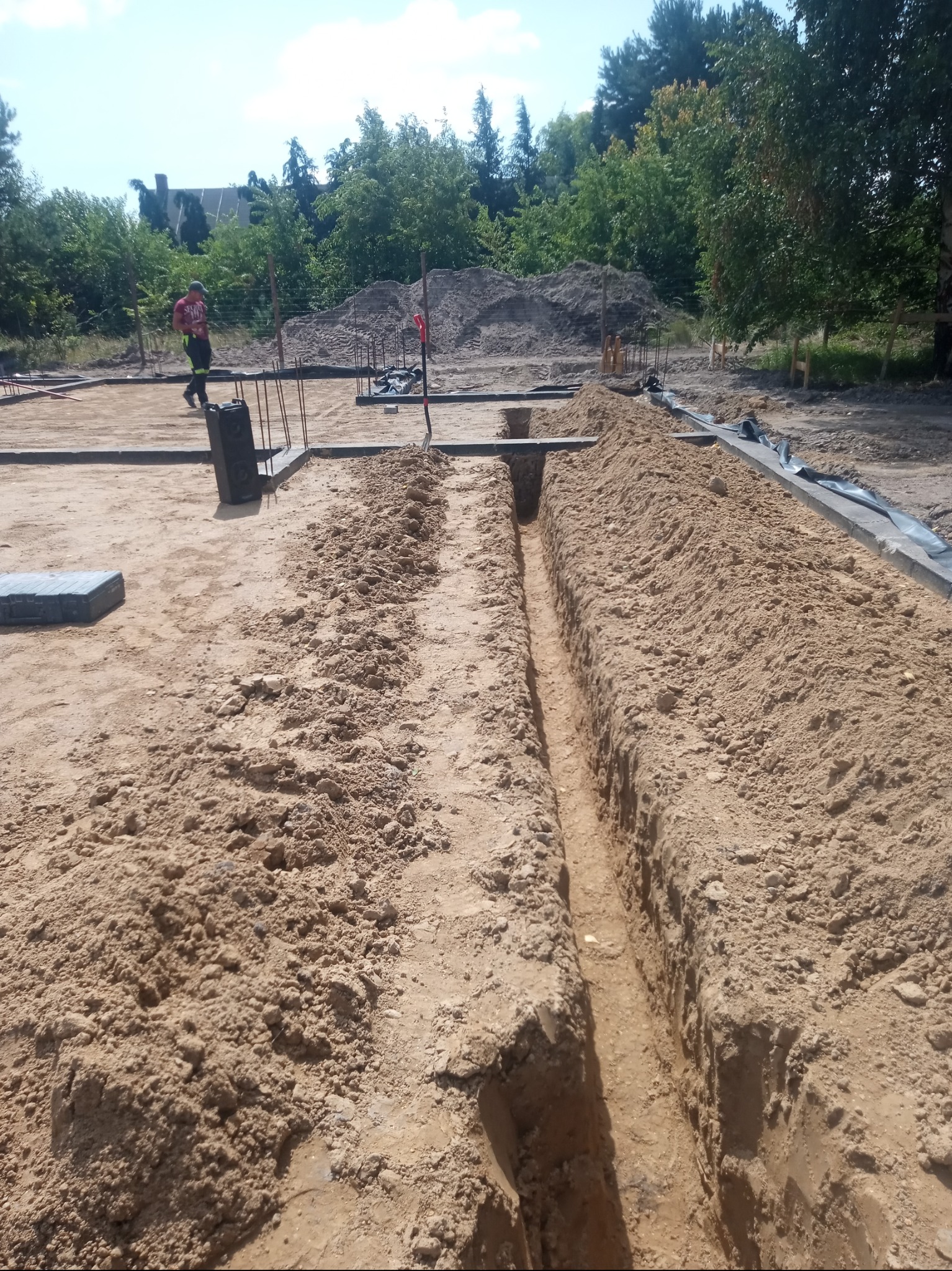 Wykop liniowy w piasku, przygotowany pod instalację, widoczny pracownik w tle i materiały budowlane.