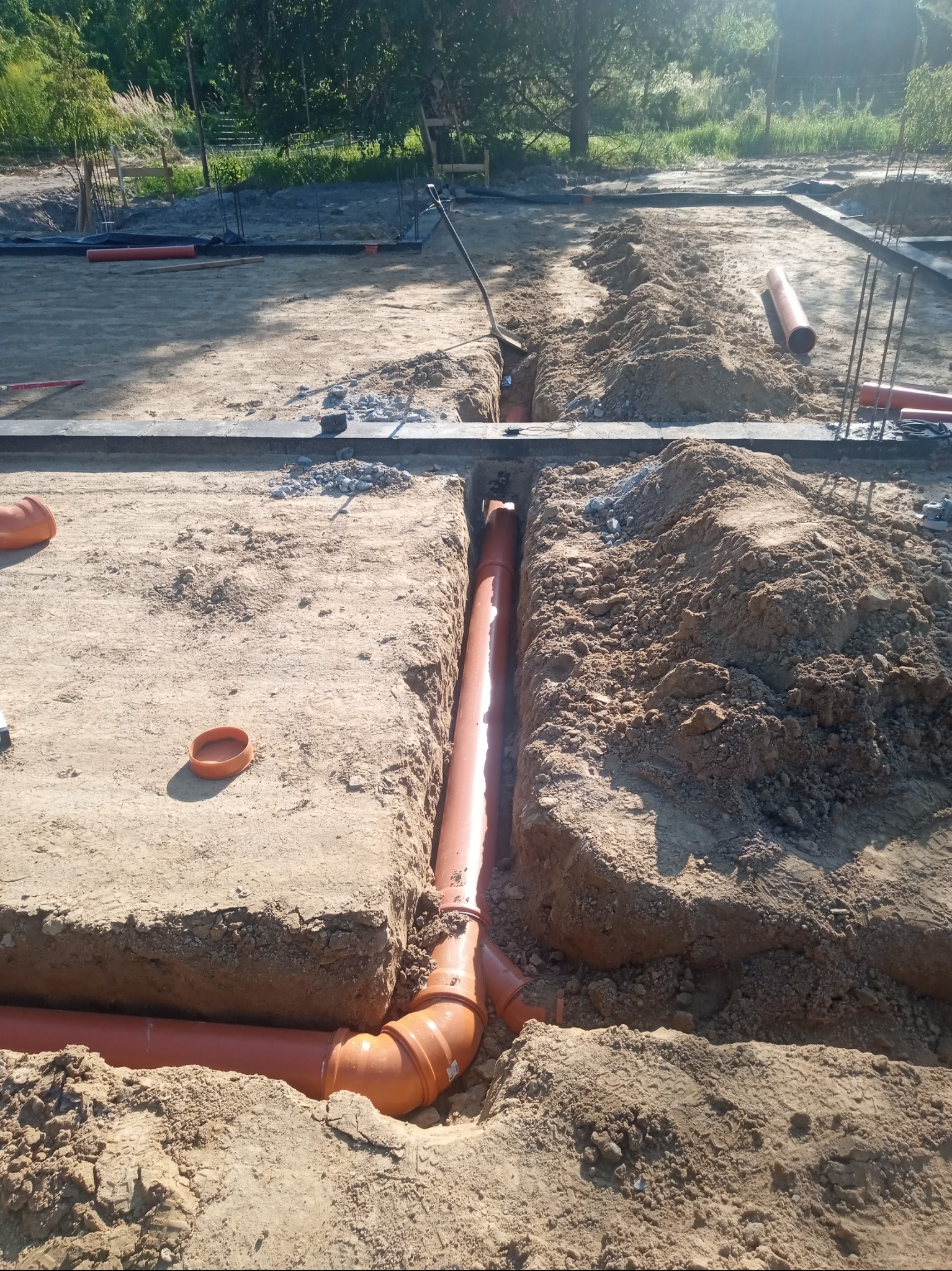 Pomarańczowe rury kanalizacyjne ułożone w wykopie w piaszczystym gruncie, widoczne złącza i fragment fundamentu budynku w tle.