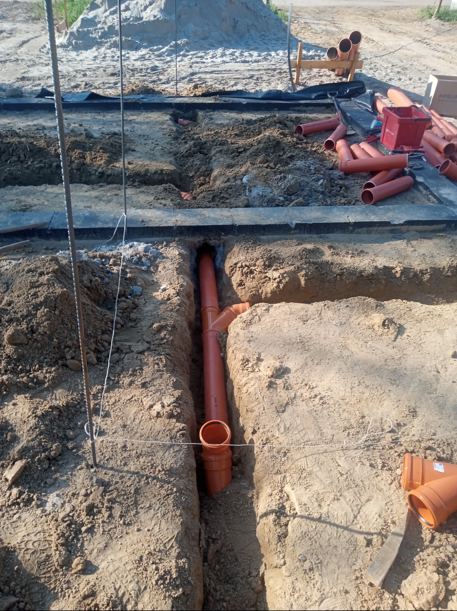 Pomarańczowe rury kanalizacyjne ułożone w wykopie ziemnym, widoczne złącza i elementy instalacji, na powierzchni ziemi leżą dodatkowe rury i narzędzia.