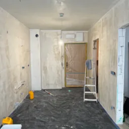 Pokój w trakcie prac przygotowawczych do wykończenia: ściany pokryte szpachlą, zabezpieczona folią podłoga, drabina oparta o drzwi zabezpieczone folią, widoczne puszki elektryczne i rury.