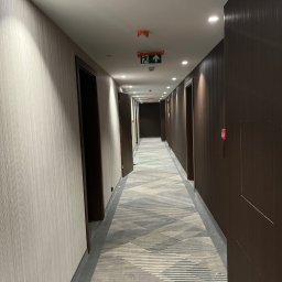 Tomasz Kobak - Długi korytarz hotelowy z tapetowanymi ścianami w odcieniach szarości i brązu, wzorzysta wykładzina podłogowa, oświetlenie sufitowe i drzwi po obu stronach.