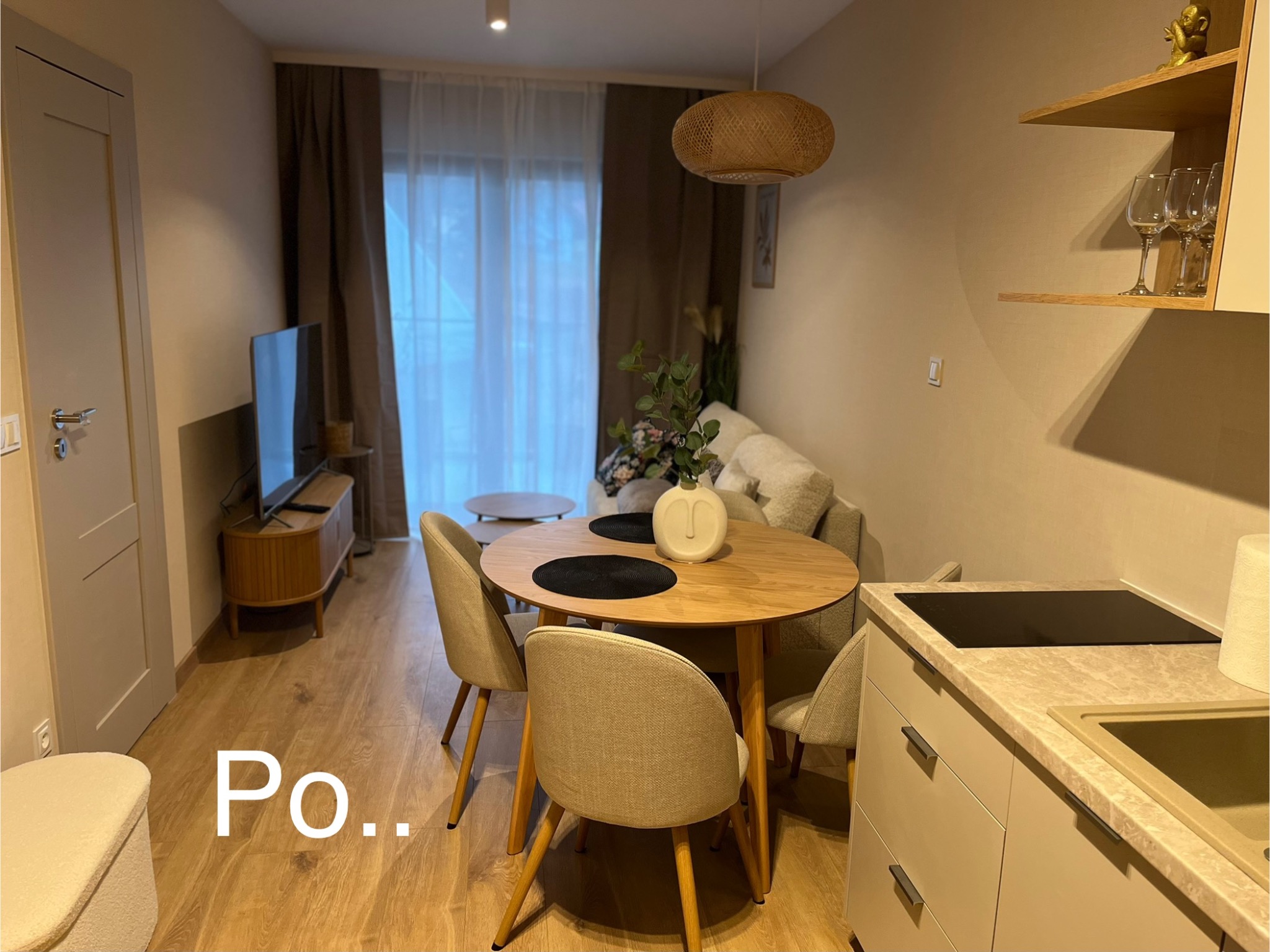 Jasne wnętrze po remoncie: okrągły stół z krzesłami, telewizor na szafce RTV, kuchnia z blatem i płytą indukcyjną, drewniana podłoga i beżowe ściany.