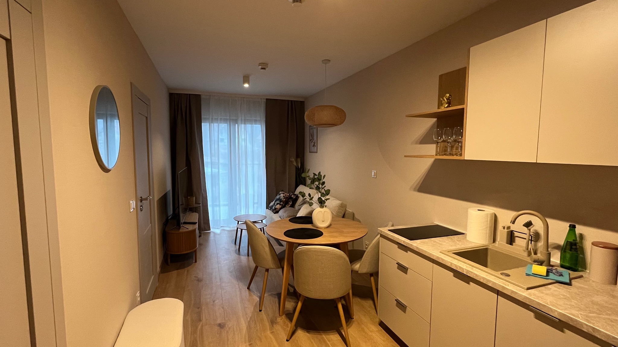 Widok na salon z aneksem kuchennym w jasnych barwach; okrągły stół jadalny z krzesłami, sofa z poduszkami w tle, minimalistyczna kuchnia z beżowymi szafkami i drewnianymi półkami.