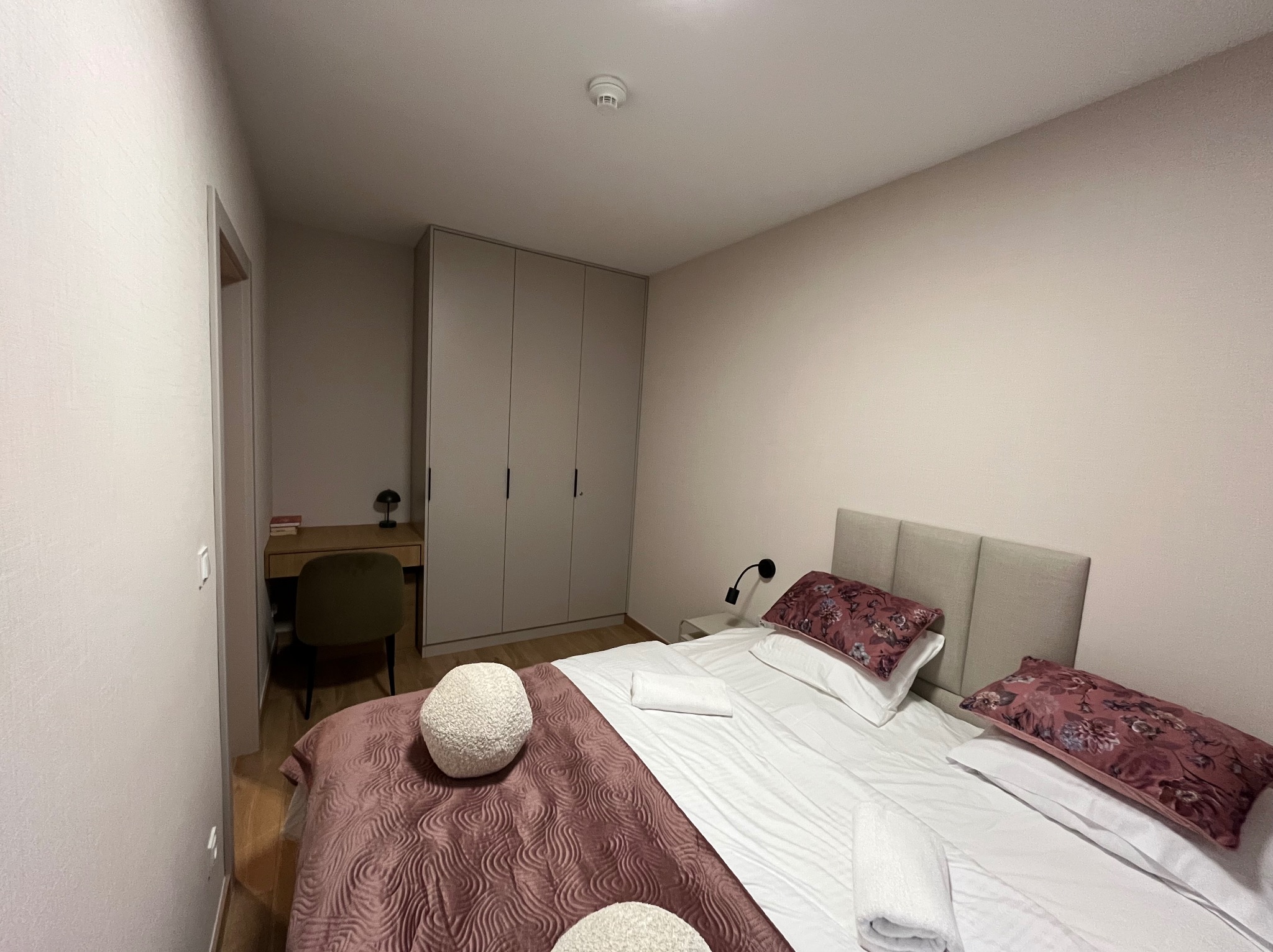 Jasna sypialnia z łóżkiem z bordową narzutą i ozdobnymi poduszkami, biurko z krzesłem w tle, ściany w odcieniach beżu.