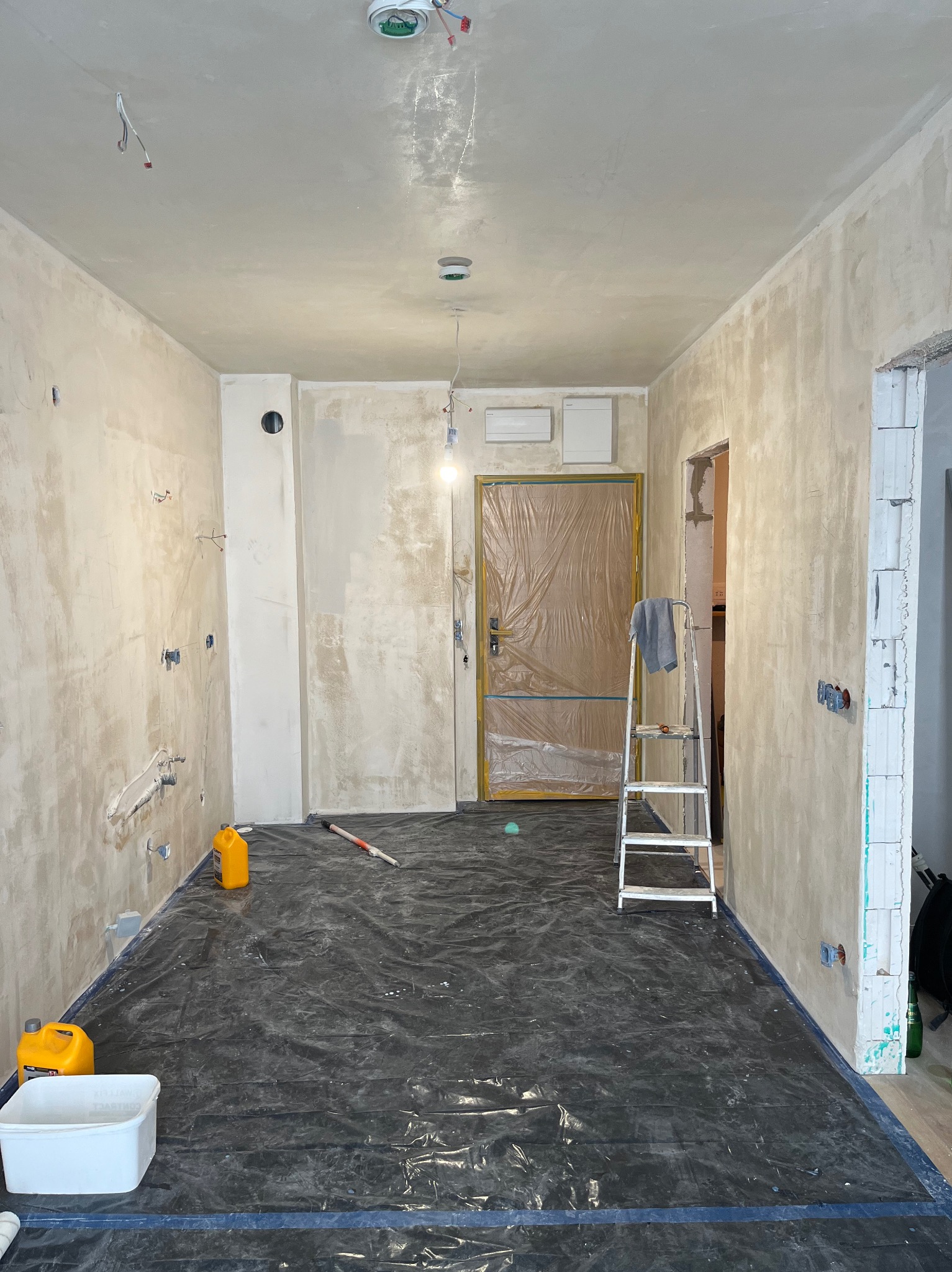 Pokój w trakcie prac przygotowawczych do wykończenia: ściany pokryte szpachlą, zabezpieczona folią podłoga, drabina oparta o drzwi zabezpieczone folią, widoczne puszki elektryczne i rury.