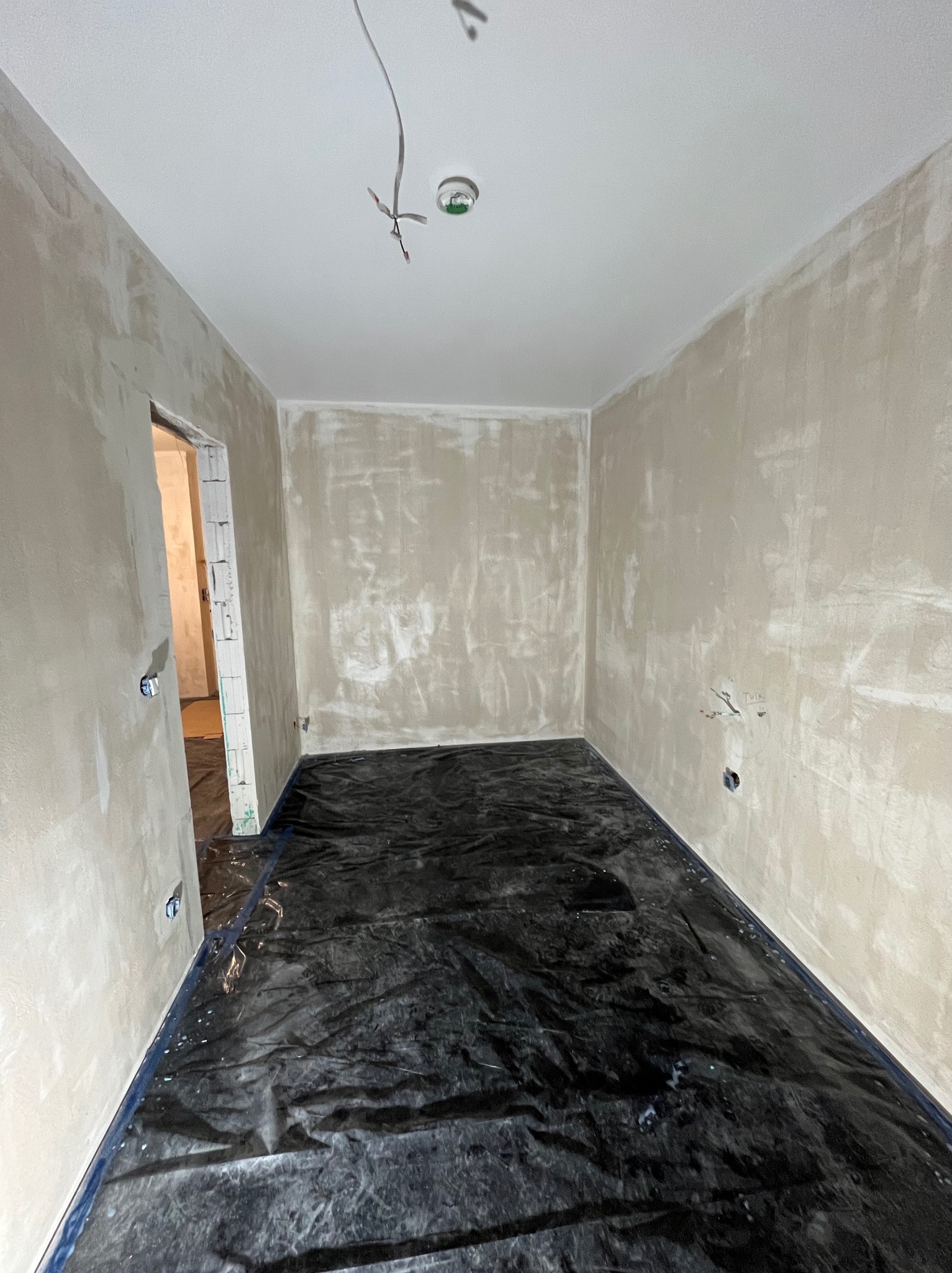 Pokój w trakcie remontu, ściany pokryte szpachlą, podłoga zabezpieczona folią malarską, widoczne wystające kable elektryczne.