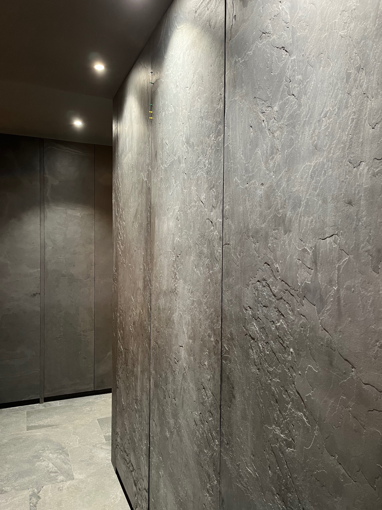 Ściana wykończona szarą, strukturalną gładzią imitującą kamień, z widocznymi w tle zabudowanymi szafami w podobnej tonacji kolorystycznej i punktowym oświetleniem sufitowym.