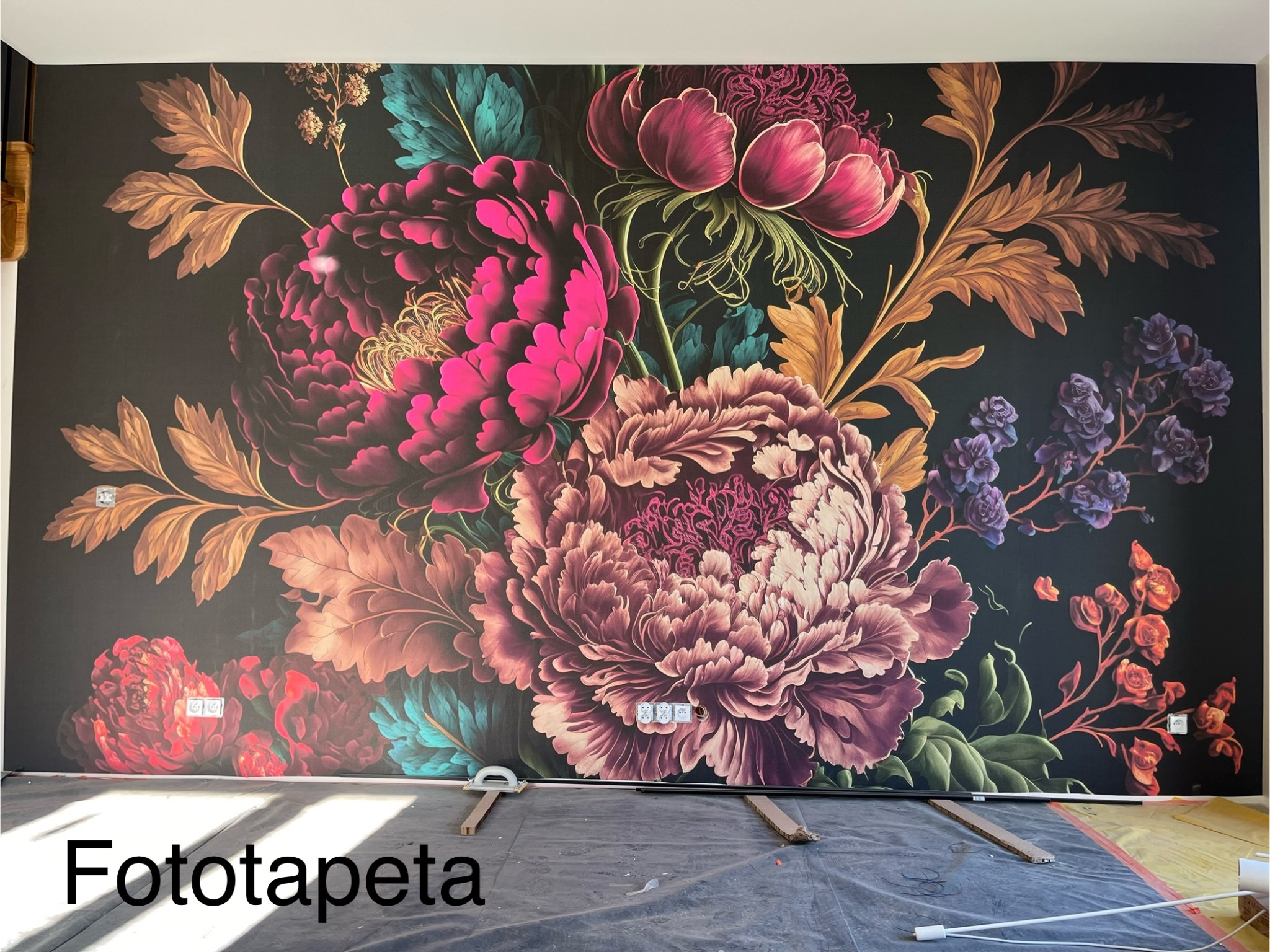 Fototapeta z motywem dużych, kolorowych kwiatów na ciemnym tle, zamontowana na ścianie w trakcie prac wykończeniowych, widoczne gniazdka elektryczne i folia ochronna na podłodze.