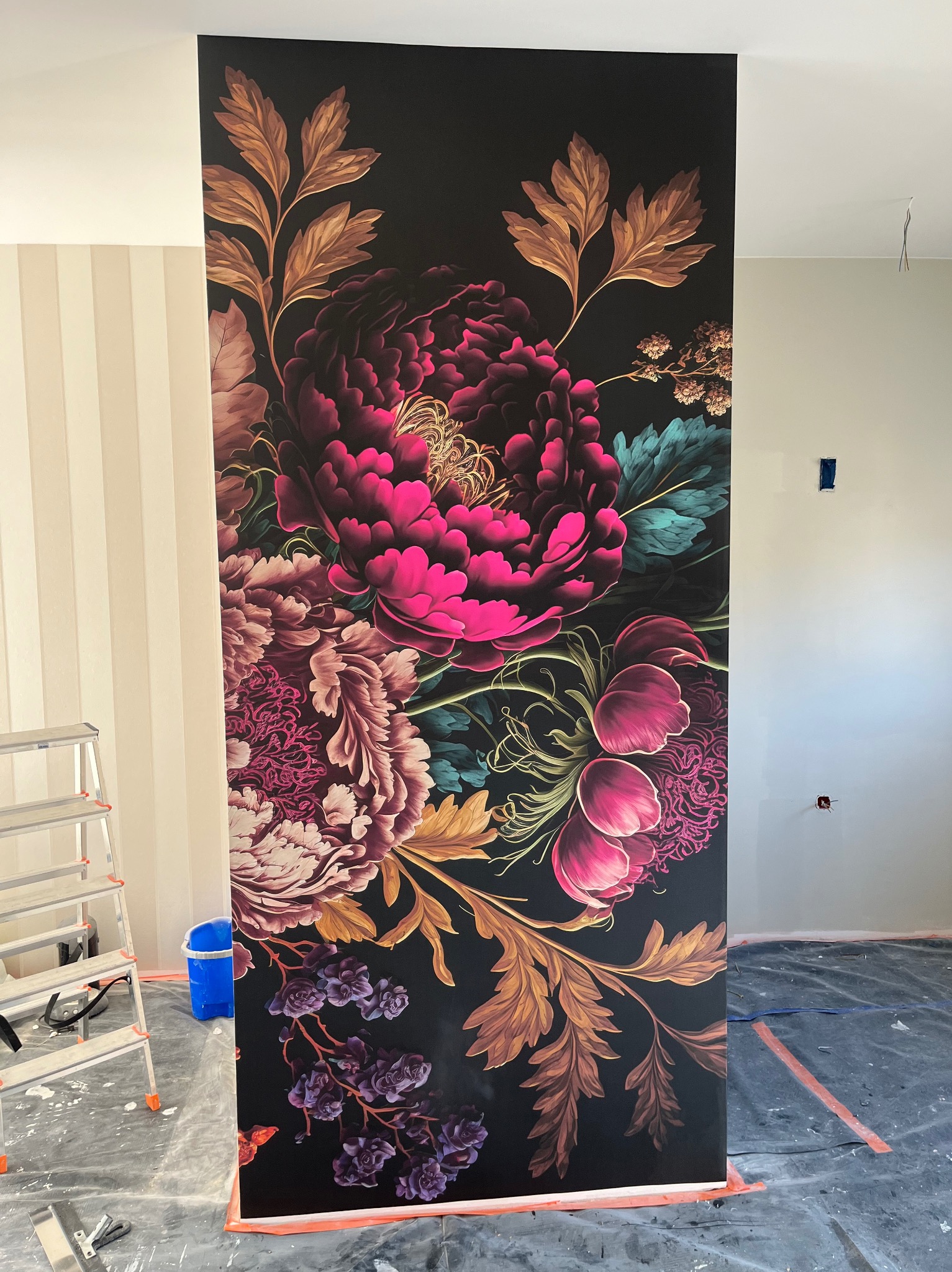 Fragment ściany z naklejoną fototapetą w motywy kwiatowe, drabina i narzędzia malarskie w tle, folia ochronna na podłodze.