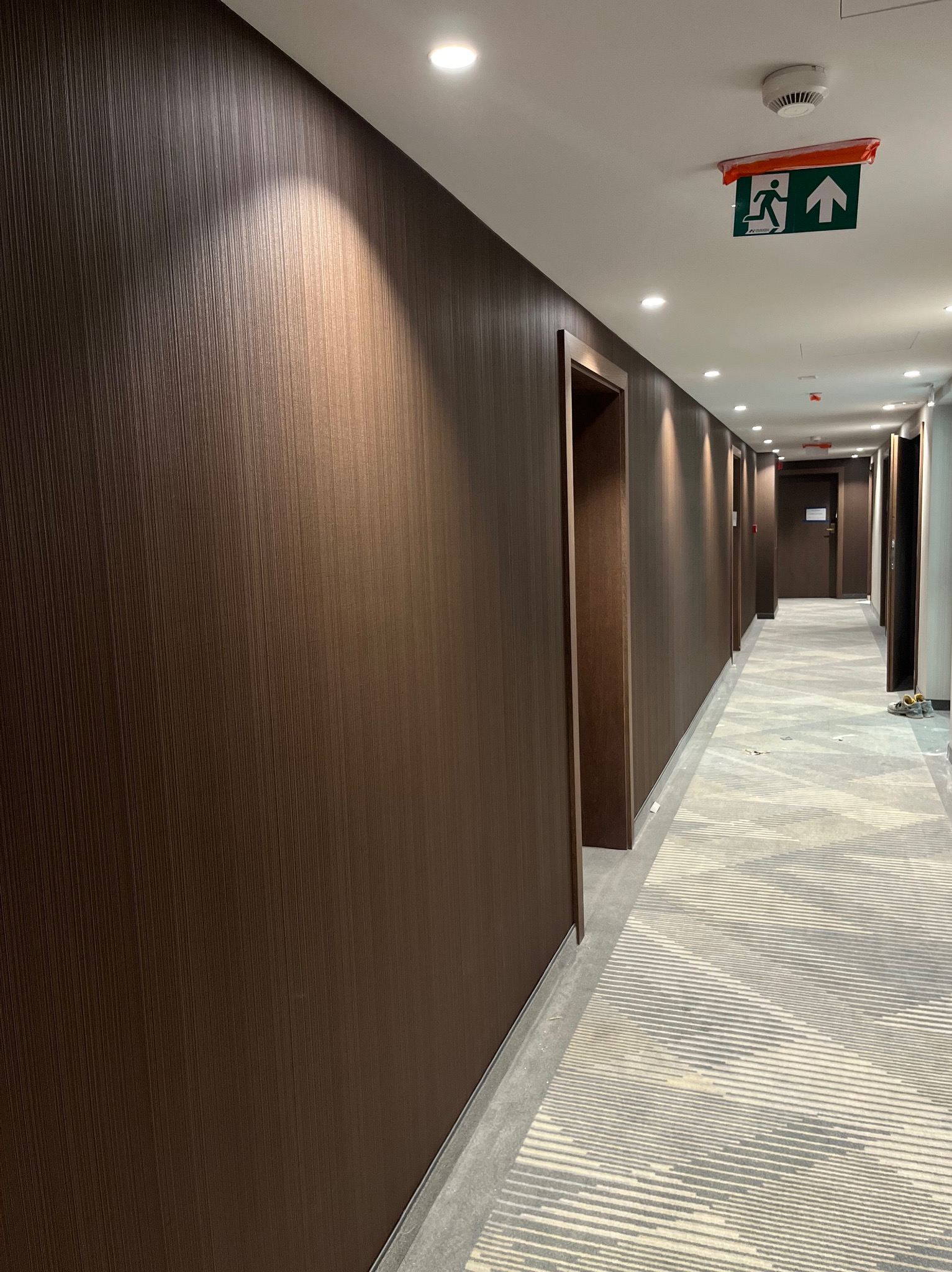 Długi korytarz w hotelu z ciemnobrązowymi ścianami o pionowej fakturze, jasnym sufitem z oświetleniem punktowym i wzorzystą wykładziną podłogową. Widoczny znak ewakuacyjny.