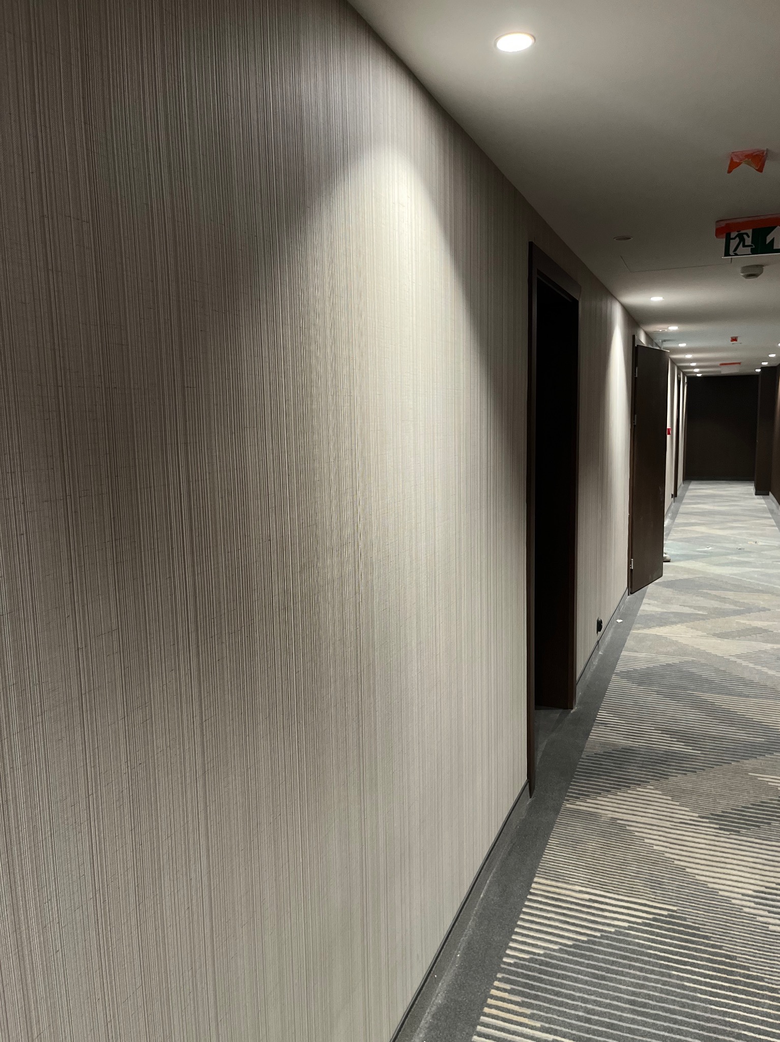 Długi korytarz hotelowy z tapetą w pionowe prążki i wzorzystą wykładziną podłogową, oświetlony punktowymi lampami sufitowymi.